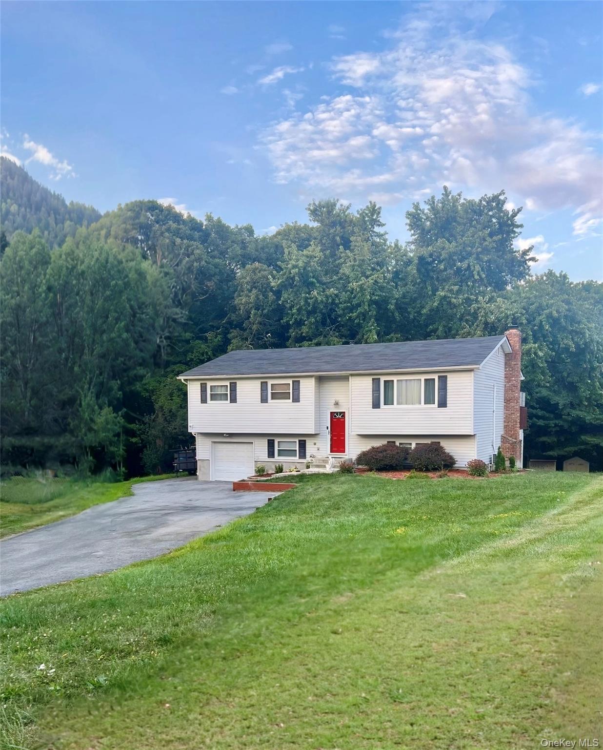 Bi-level home with driveway, an attached garage, a front lawn, a chimney, and a wooded view