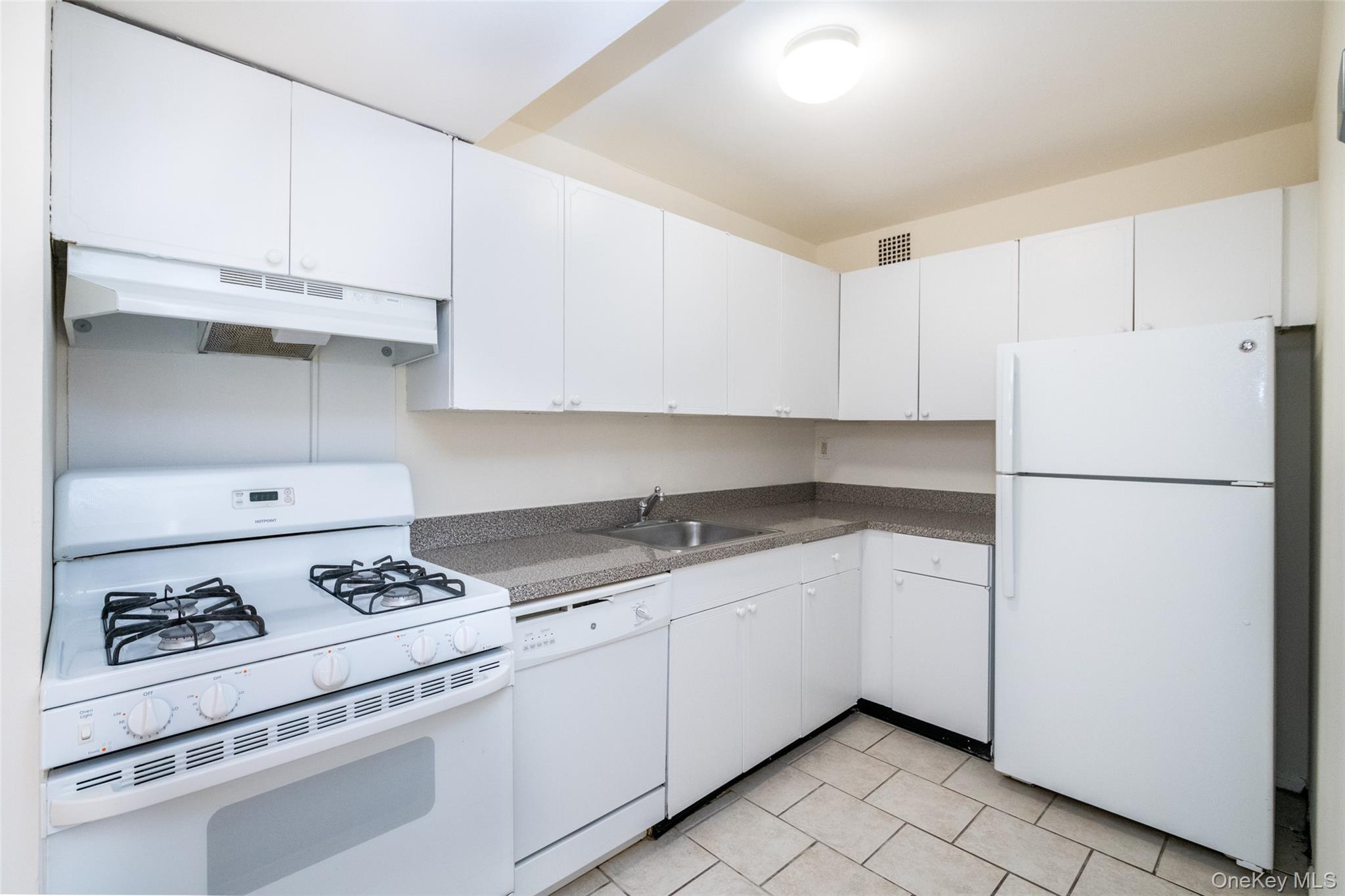Kitchen with white appliances, under cabinet range hood, white cabinets, dark countertops, and light tile patterned flooring