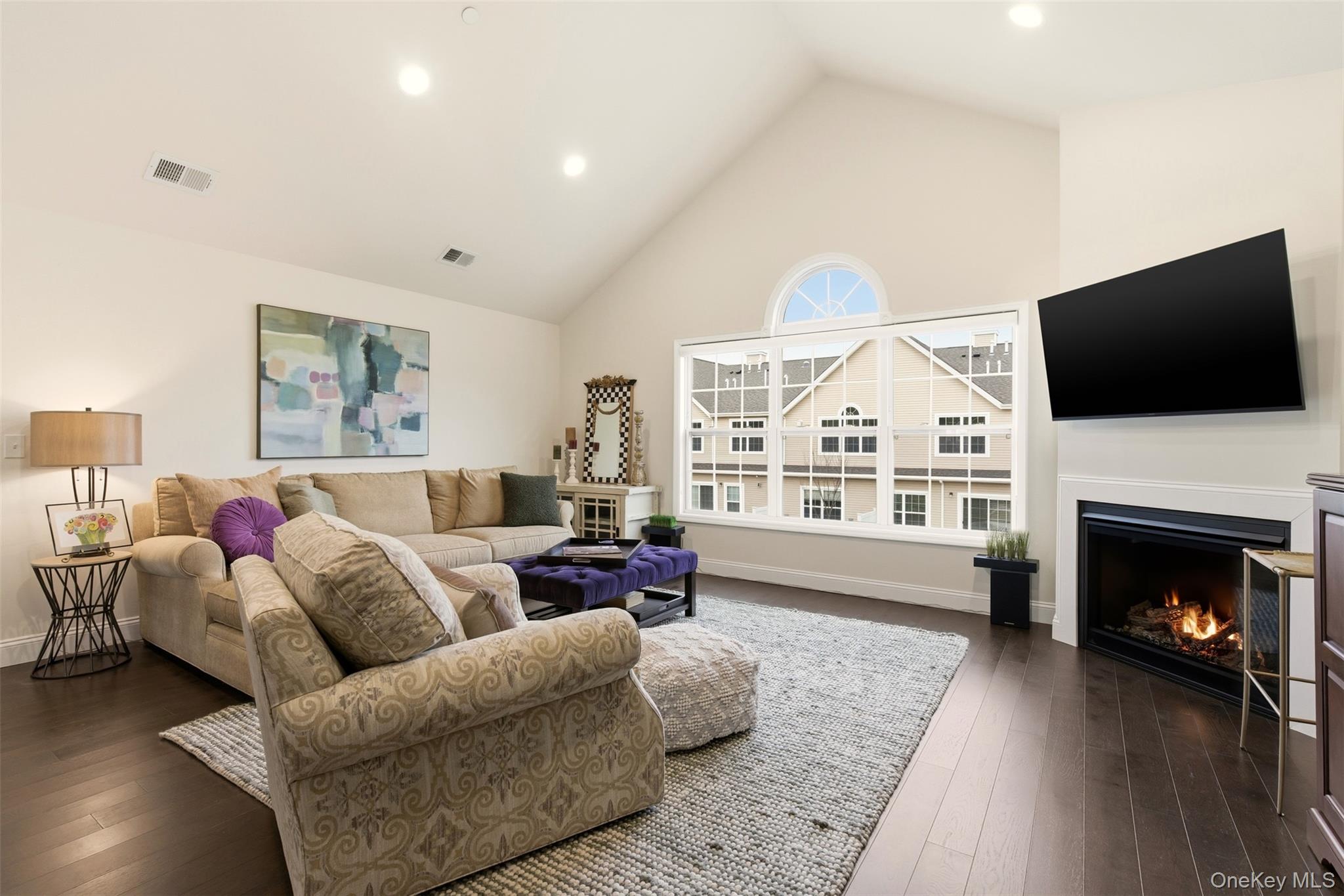 Living area with a warm lit fireplace, high vaulted ceiling, dark wood-style floors, and recessed lighting