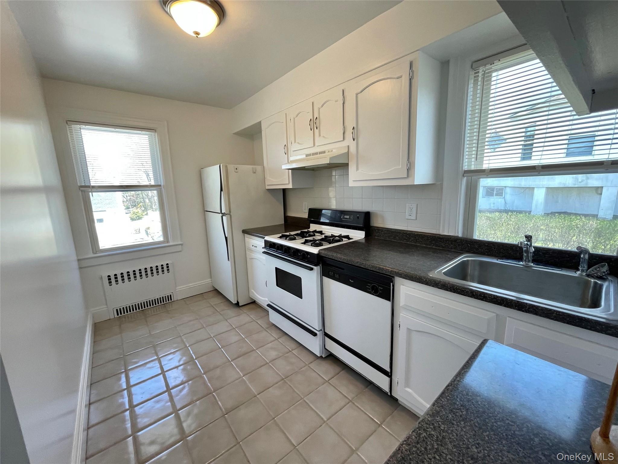 Kitchen with radiator heating unit, under cabinet range hood, dark countertops, a sink, and white appliances
